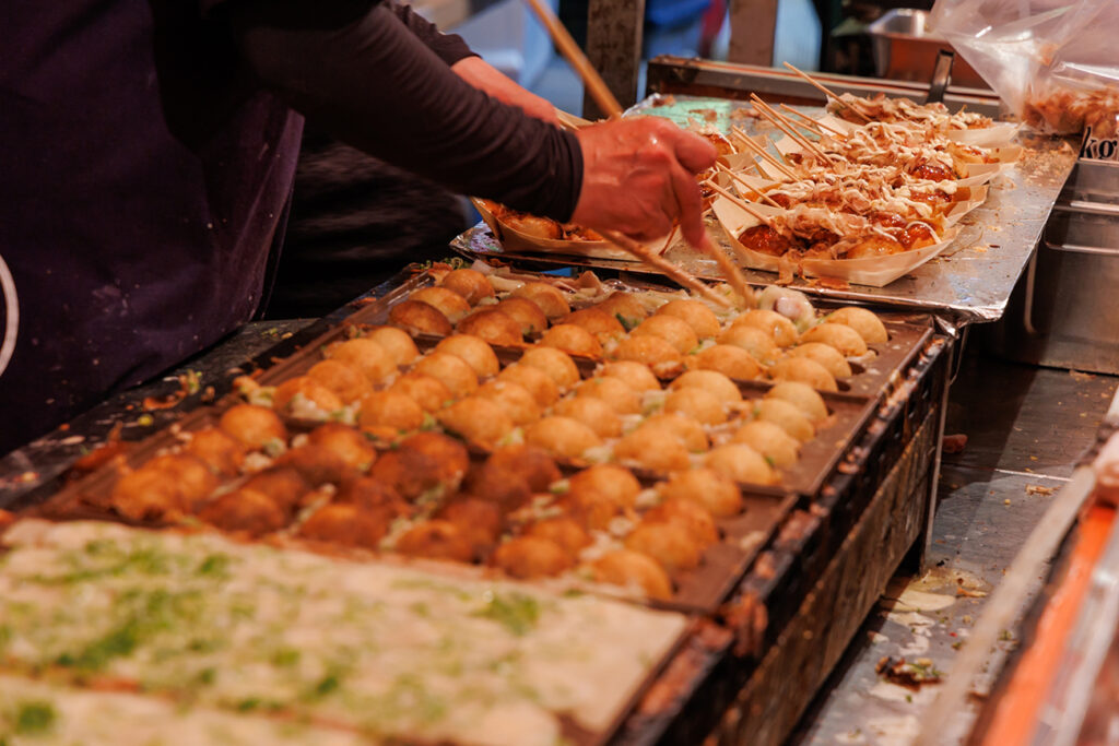 Experience the vibrant culture of Japan through this captivating image of Takoyaki, a popular street food, being prepared at a festival.