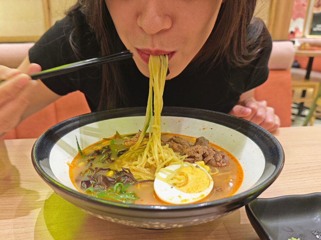 Close-up of a woman enjoying a delicious bowl of ramen with chopsticks, featuring a rich broth, noodles, sliced egg, and tender meat in a cozy restaurant setting